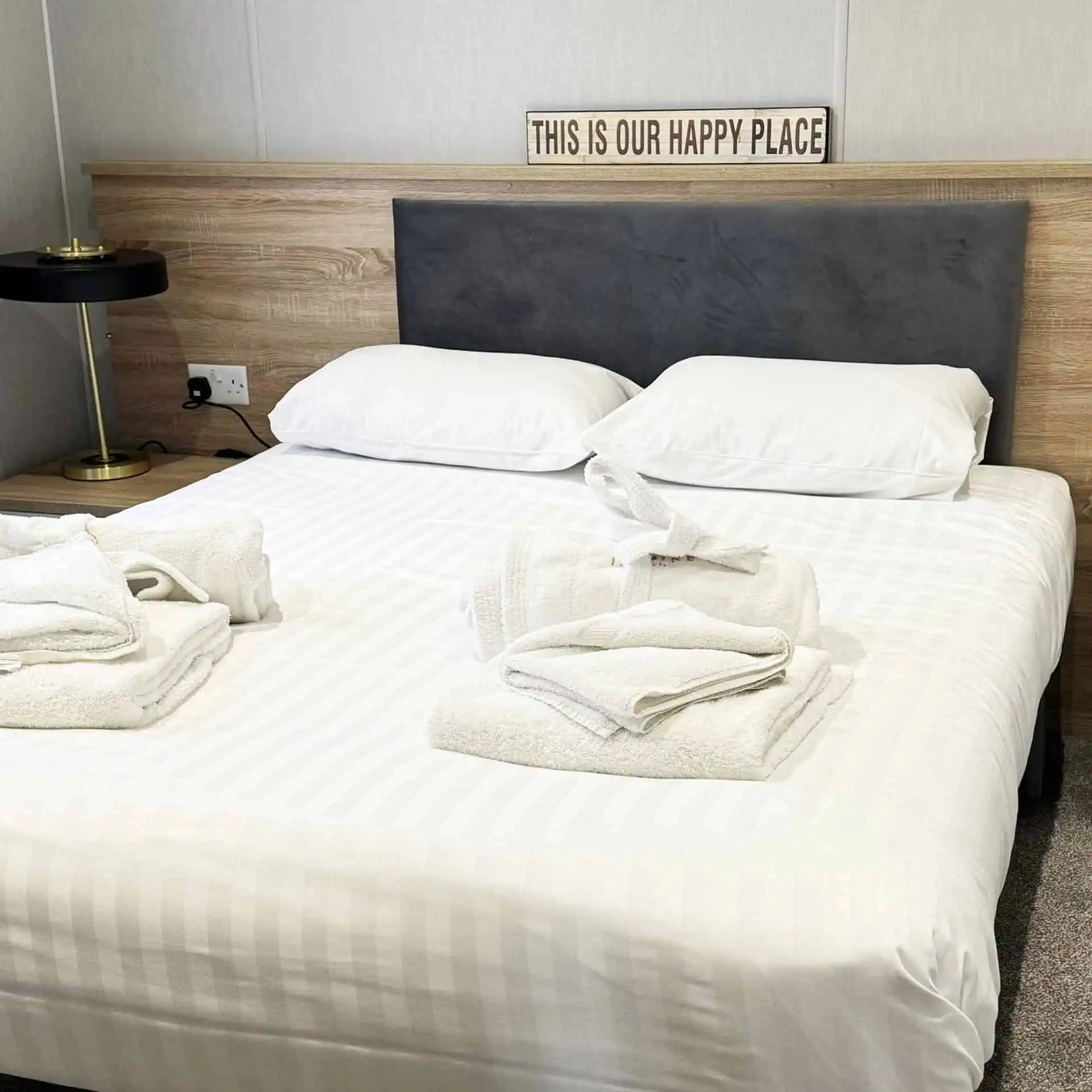 A made bed with white striped bedding and fresh towels, and a wooden sign above reading "THIS IS OUR HAPPY PLACE" in a holiday home bedroom - Club4.