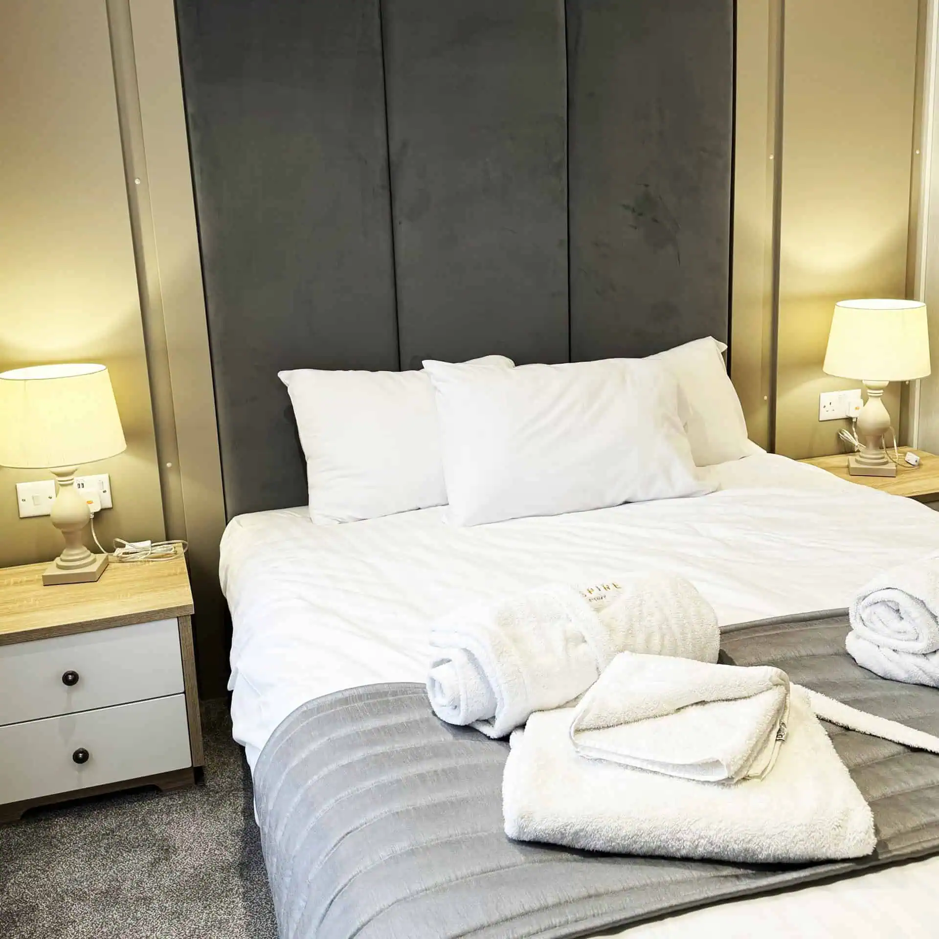 A bedroom with fresh white linens and towels on a bed with a tall gray headboard by bedside tables with matching table lamps with warm lighting - Club4.