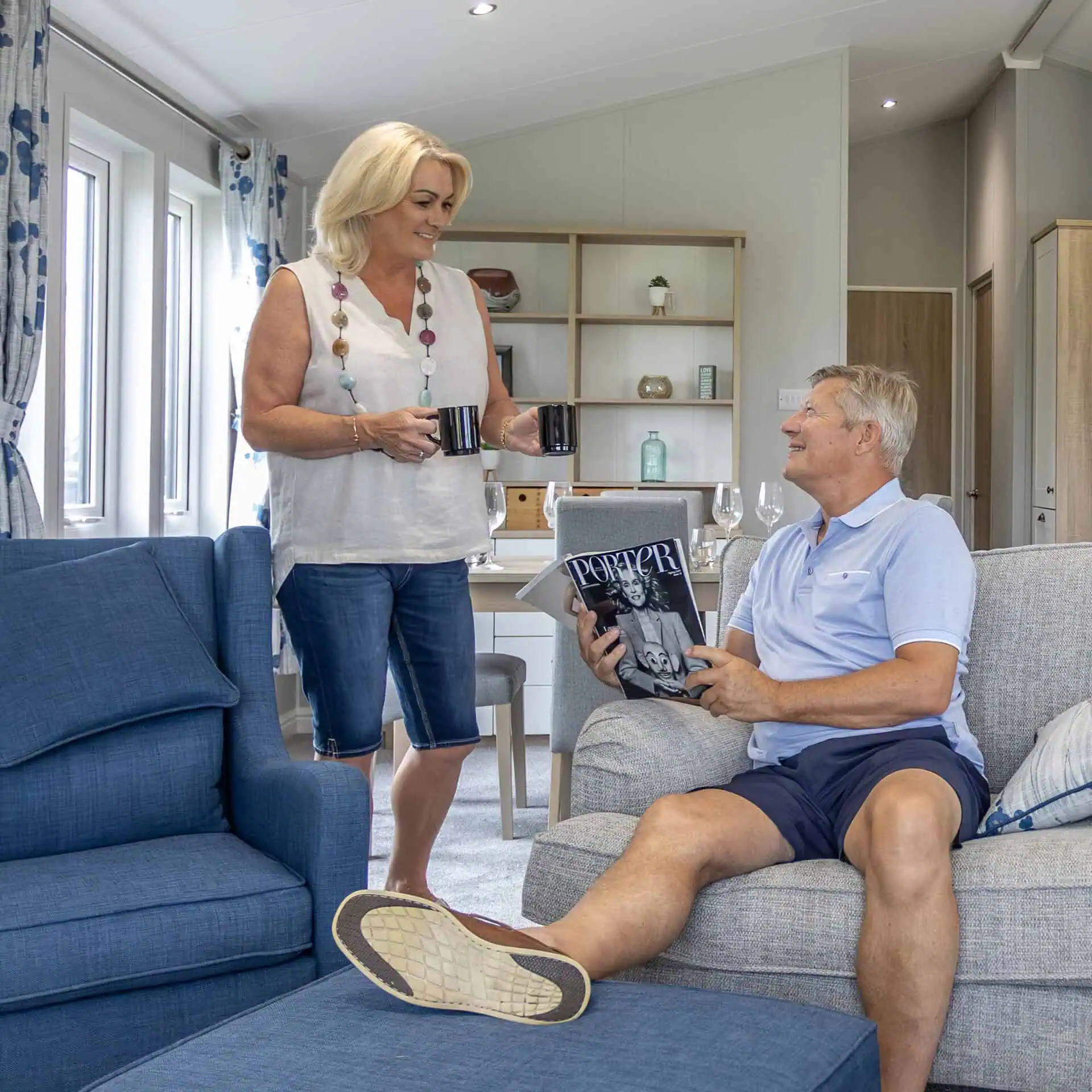 A couple sharing coffee in a holiday home living room, one standing with coffee mugs while the other sits on a gray sofa reading Porter magazine - Club4.