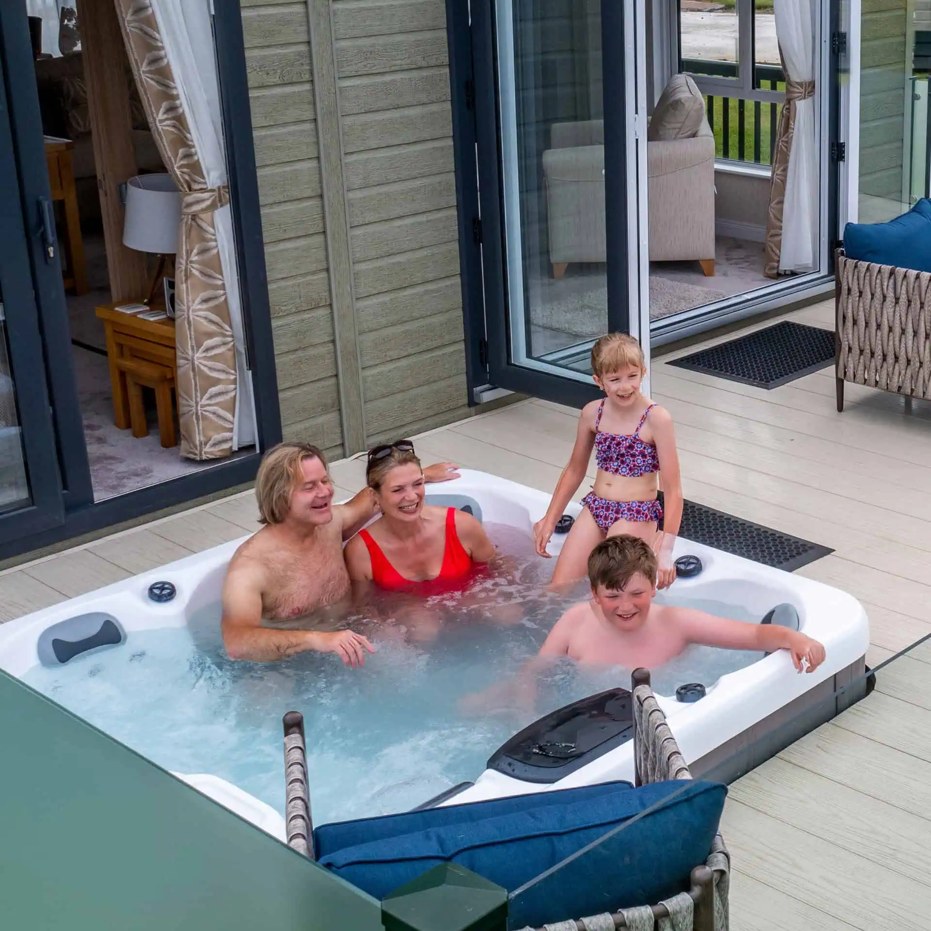 A family enjoying a hot tub on a deck outside their holiday home - two adults and two children laughing and playing in the bubbling water - Club4.