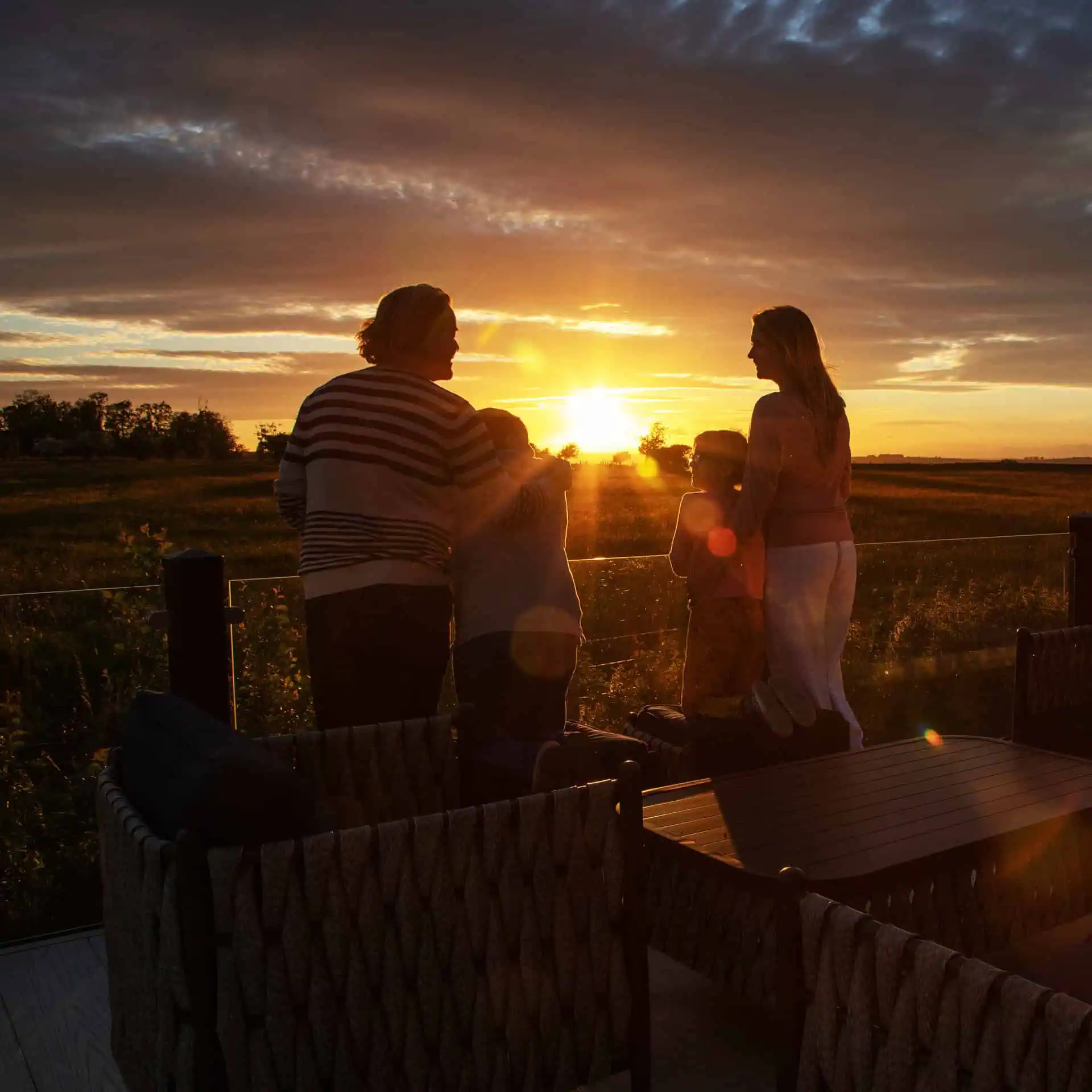 Silhouetted family watching the sunset from a deck, two adults and two children standing together against an orange and gold sky over rural fields - Club4.