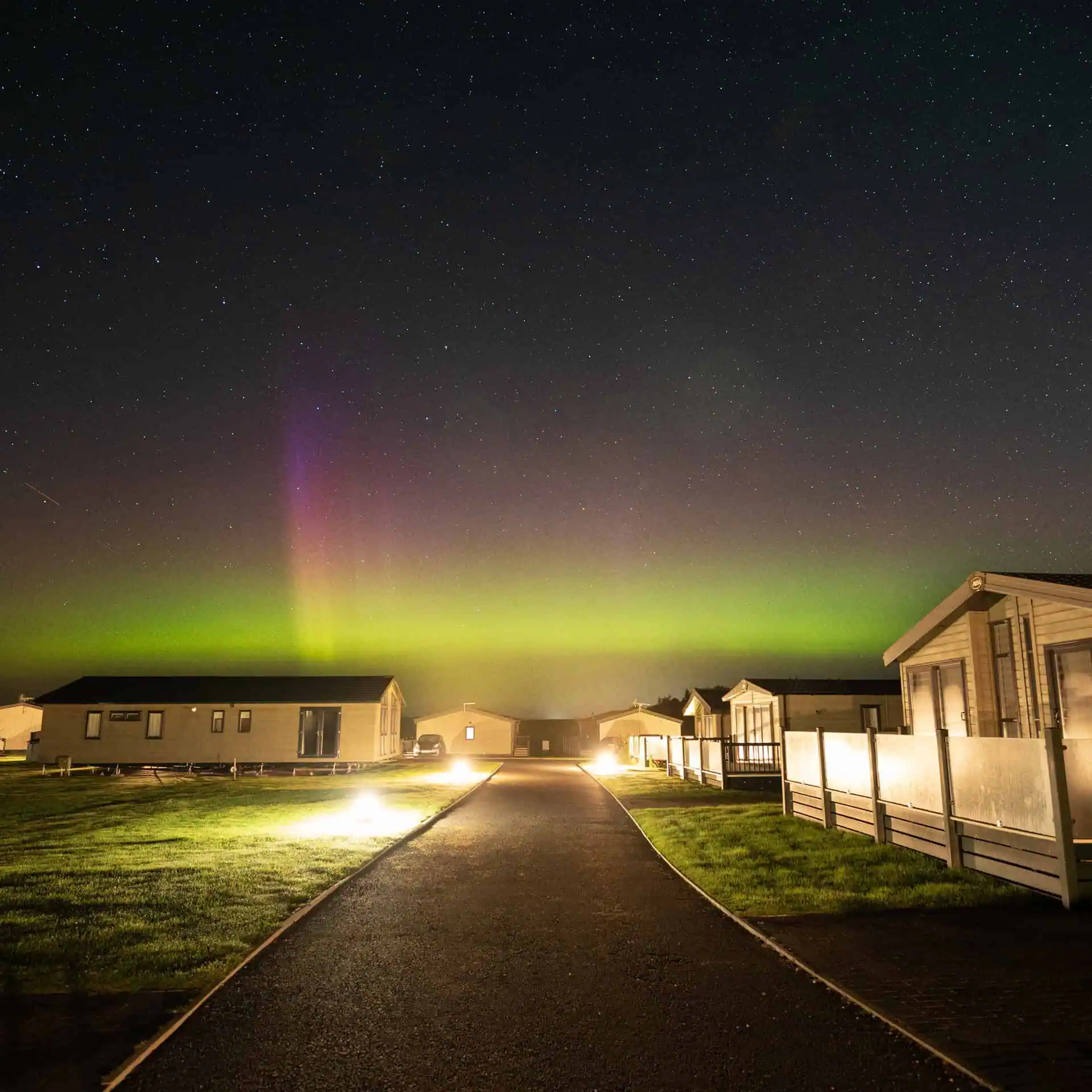 The Northern Lights in the night sky above a holiday park, illuminated caravans and a lit pathway - Club4.