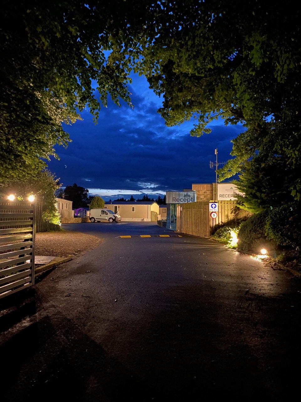 Caravan park entrance at night, framed by trees with a view of the reception area in the distance.