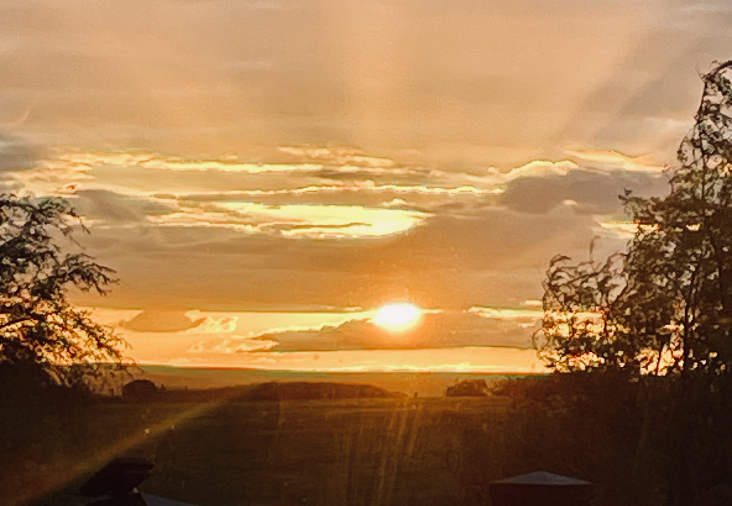 Bright orange sunset with sun rays peeking through clouds over a countryside landscape.