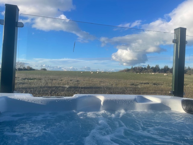 A bubbling hot tub with a clear view of a grassy field and blue sky beyond glass fencing.