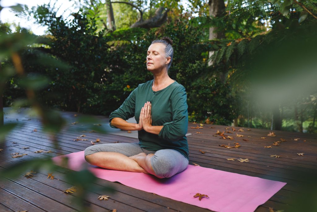 Senior woman sitting cross-legged meditating on pink yoga mat on deck in garden with autumn leaves Senior woman sitting cross-legged meditating on pink yoga mat on deck in garden with autumn leaves