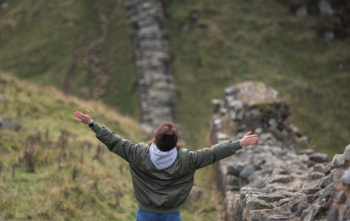 Person standing with arms outstretched on a countryside path beside a stone wall, representing freedom, independence, and solo travel in nature.